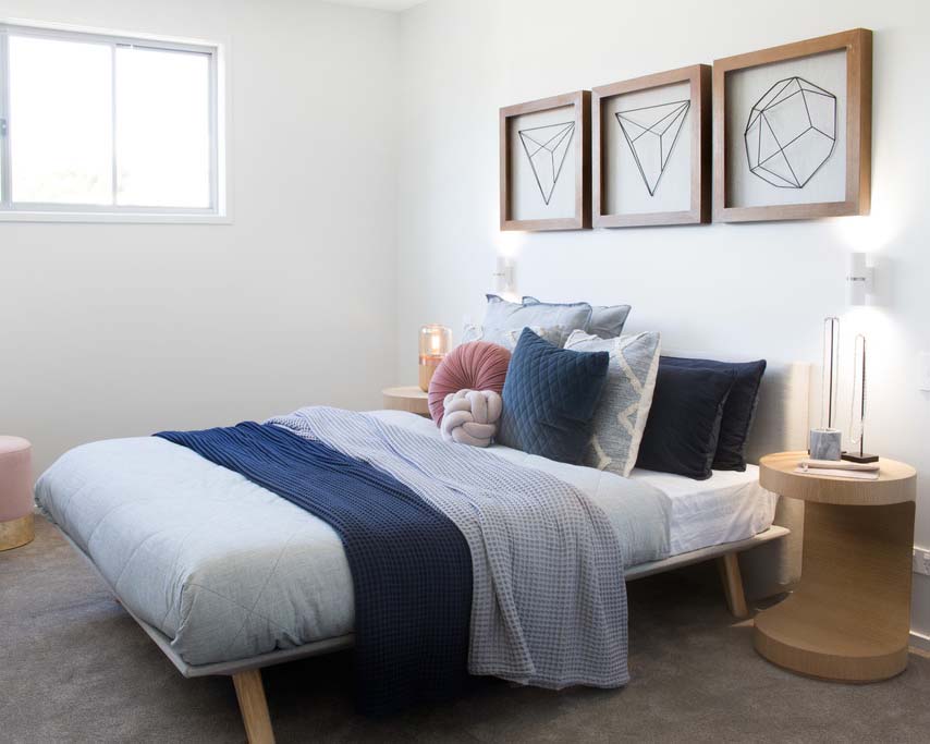 A double bed dressed with blue grey and pink bedding and accessories and a unique wooden bedside table.