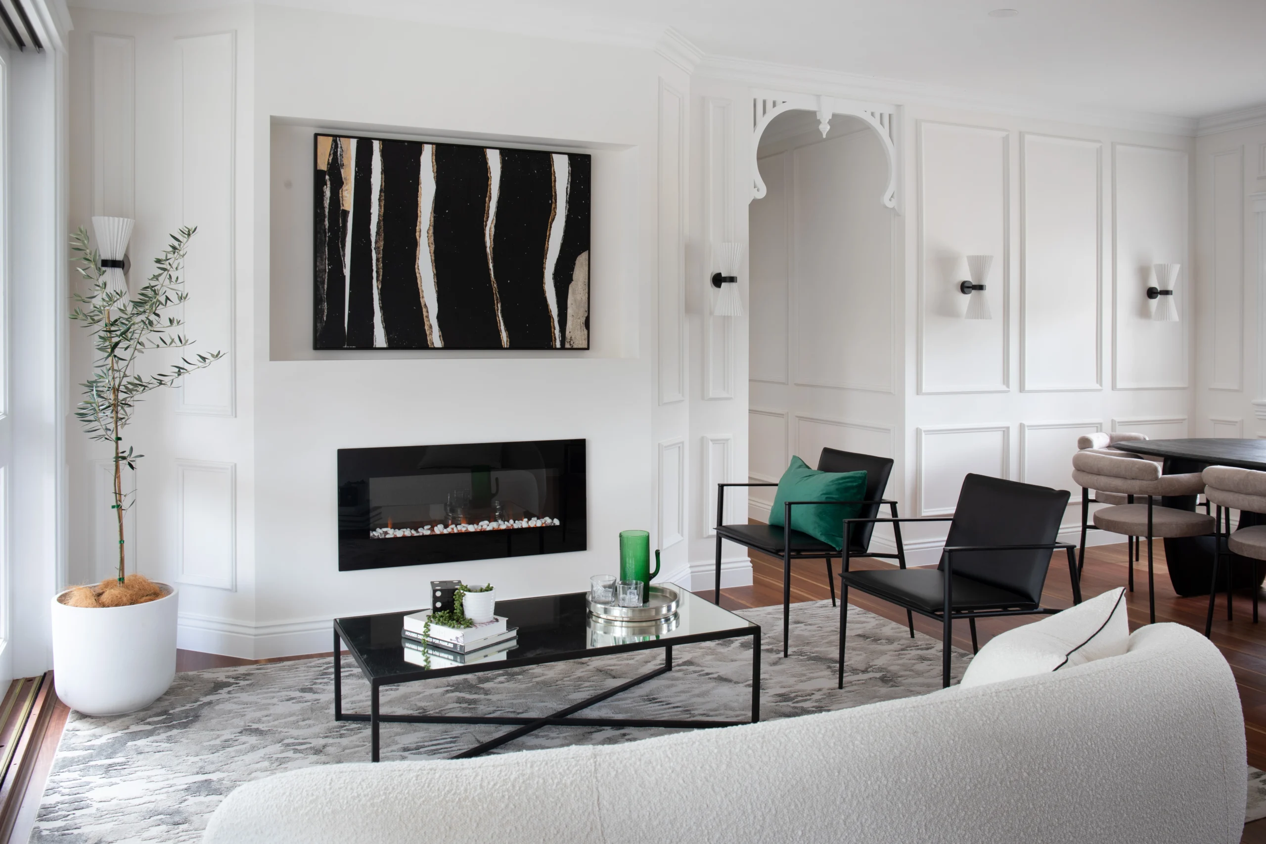 Modern living room in a beautifully styled home featuring soft neutral tones, black accent chairs, a glass coffee table, and green decor touches. Wall panelling, artwork, and a fireplace add elegance.