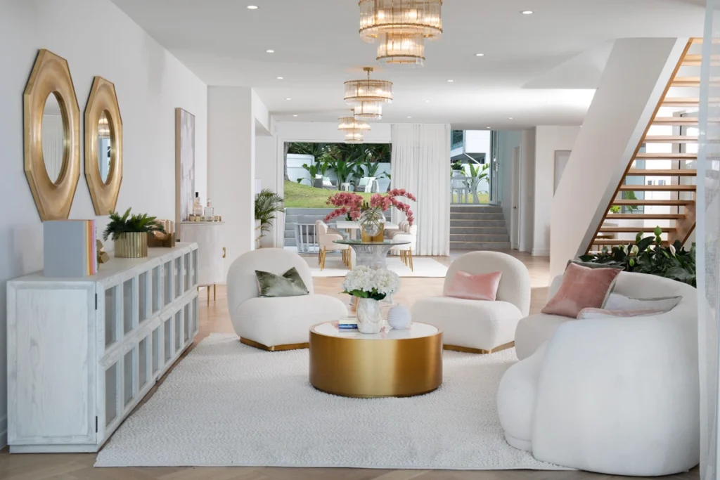 Elegant living and dining area showcasing expert property styling with soft white boucle chairs, gold accents, a round coffee table, and pink decor touches. Stylish chandeliers and large mirrors enhance the bright, open space designed for luxury living.