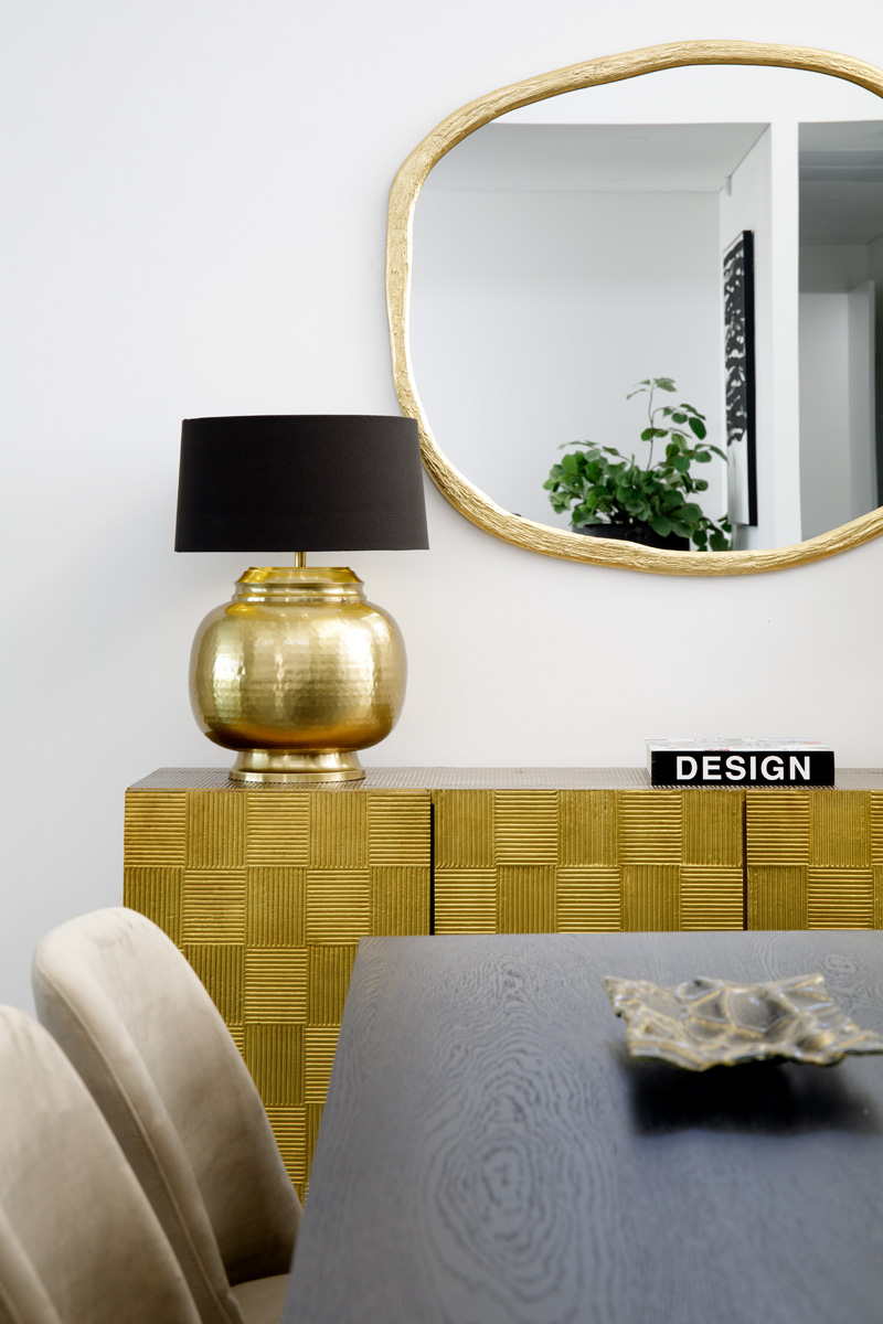 A dining area with a gold sideboard styled with a lamp and gold framed mirror