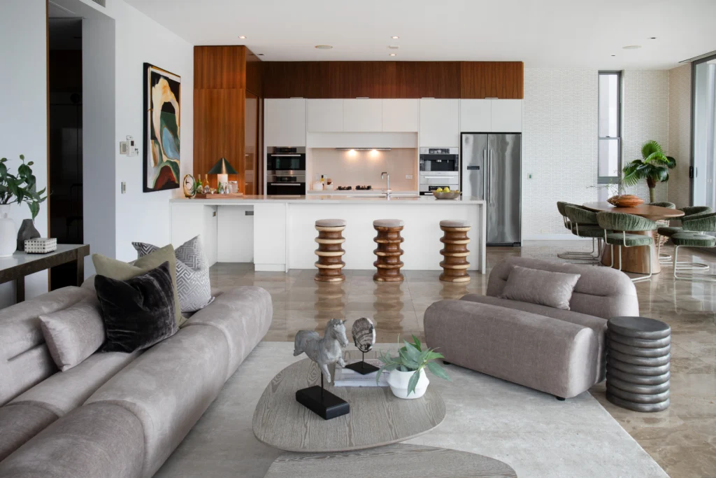 Modern open-plan living, kitchen, and dining area featuring neutral-toned furniture, sculptural bar stools, marble floors, and green velvet dining chairs. Styled with natural textures and contemporary decor.”