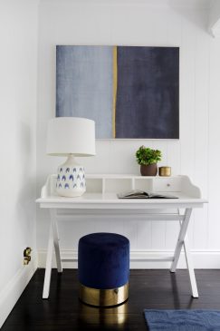 A white desk staged under a large piece of wall are with a blue and gold stool and a large lamp